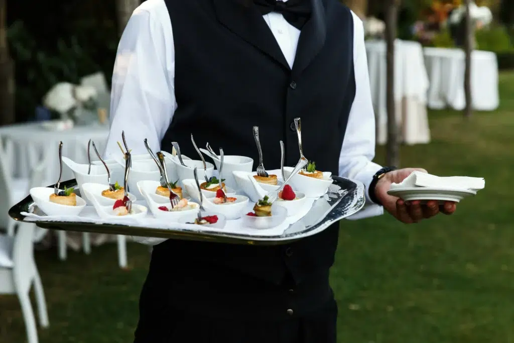 Serveur en tenue élégante portant un plateau de bouchées raffinées lors d’un service traiteur en plein air.